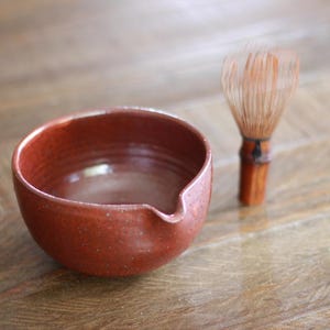 May include: A terracotta-colored ceramic bowl with a small pouring spout and a speckled finish, accompanied by a light brown bamboo matcha whisk featuring thin, delicate tines and a central dark binding cord on the handle.
