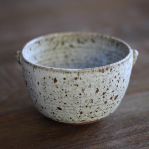 Peut inclure: Un bol en céramique moucheté avec deux petites anses. Le bol est blanc cassé avec des mouchetures marron et une texture légèrement rugueuse. Le bol est posé sur une surface en bois.