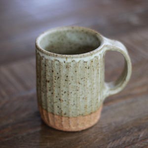 May include: A cylindrical ceramic mug featuring a speckled light green glaze on the upper portion and a textured, unglazed terracotta-colored base. The exterior surface has vertical fluted grooves and a single curved handle on the right side.