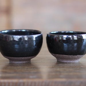May include: Two small, black ceramic bowls with a glossy finish. The bowls have a rounded shape and sit on a slightly textured, brown base. The interior of the bowls reflects light, creating highlights.
