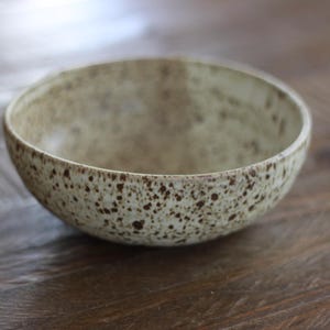 May include: A round ceramic bowl with a cream-colored base and a speckled pattern of dark brown spots across the exterior and interior surfaces. The bowl features a smooth, curved rim and a deep, open basin, showcasing a consistent speckled texture.