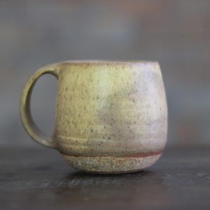 May include: A handmade ceramic mug with a rounded body and a sturdy handle. The mug has a speckled, earthy tone with a light brown base and a darker rim. The mug is designed for hot beverages.