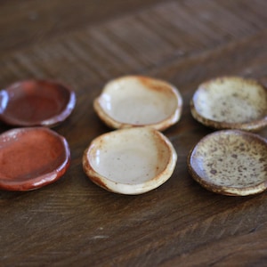 May include: Six small, handmade ceramic dishes in shades of brown and beige. The dishes have a shallow, rounded shape and a rustic, speckled glaze. They are arranged on a wooden surface, showcasing their unique textures and colors.
