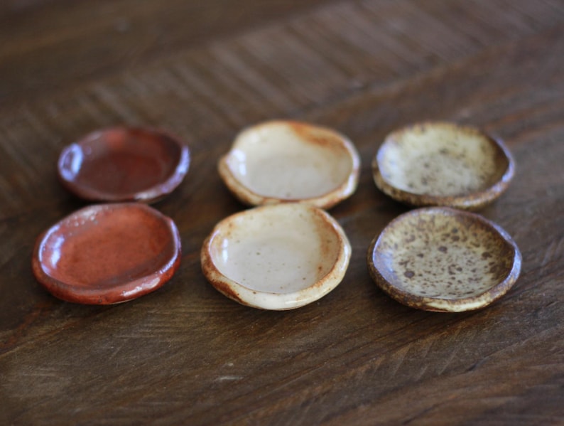 May include: Six small, handmade ceramic dishes in shades of brown and beige. The dishes have a shallow, rounded shape and a rustic, speckled glaze. They are arranged on a wooden surface, showcasing their unique textures and colors.
