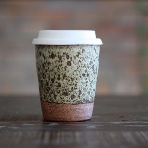 May include: A ceramic travel mug with a white lid. The mug has a speckled green and brown pattern on the upper portion and a brown base. The mug is designed for hot or cold beverages.