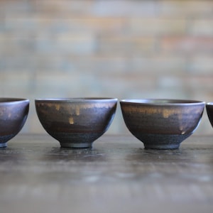 May include: A set of four ceramic bowls with a dark brown and black exterior finish. Each bowl features a rounded shape with a slightly flared rim and a distinct drip-like glaze pattern in a lighter tan color near the top edge of the vessel.