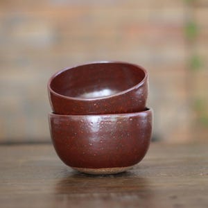 May include: Two stacked, handmade, reddish-brown ceramic bowls. The bowls have a rounded shape with a slightly textured surface and a natural, unglazed base. The interior of the bowls is a glossy, darker shade of brown.