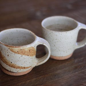 May include: Two ceramic mugs with speckled off-white glaze and a rounded handle. The front mug features a swirling brown pattern, while the back mug is plain speckled off-white. Both mugs have an exposed, unglazed terracotta-colored base.