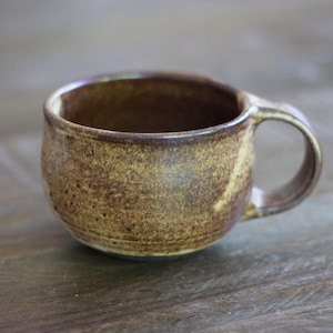 May include: A handmade, small ceramic mug with a rounded body and a single handle. The mug has a speckled, earthy brown glaze with darker brown accents. The interior is a darker brown color.