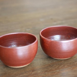 May include: Two small, round, reddish-brown ceramic bowls. The bowls have a smooth, glossy finish and a simple, minimalist design. They are sitting on a wooden surface.