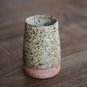 May include: A cylindrical ceramic tumbler featuring a speckled light green and brown upper section and a solid, textured reddish-brown base. The interior of the vessel is light-colored and speckled, showing a smooth rim and a tapered body shape.