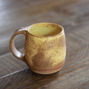 May include: A ceramic mug with a rounded body and a curved handle. The exterior features a speckled yellow glaze that transitions into a warm brown tone at the base. The interior of the mug is a smooth, light yellow finish with visible dark speckles.