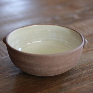May include: A round ceramic bowl with a textured, matte brown exterior and a smooth, glossy cream-colored interior. The bowl features two small, rounded handles on opposite sides, providing a simple and functional design for serving food.