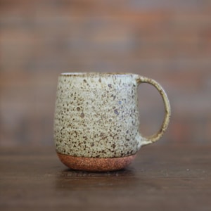 May include: A speckled ceramic mug with a rounded base and a large handle. The mug has a light beige color with brown speckles and a reddish-brown base. The mug is designed for hot beverages.