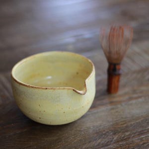 May include: A pale yellow ceramic bowl with a small pouring spout and a speckled finish, accompanied by a brown bamboo matcha whisk. The bowl has a rounded shape and a smooth interior, while the whisk features thin, delicate bristles.