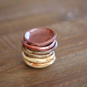 May include: A stack of small, handmade ceramic dishes in varying earth tones. The top dish is a deep reddish-brown, followed by shades of brown and beige. The dishes are stacked on top of each other, creating a rustic and natural aesthetic.