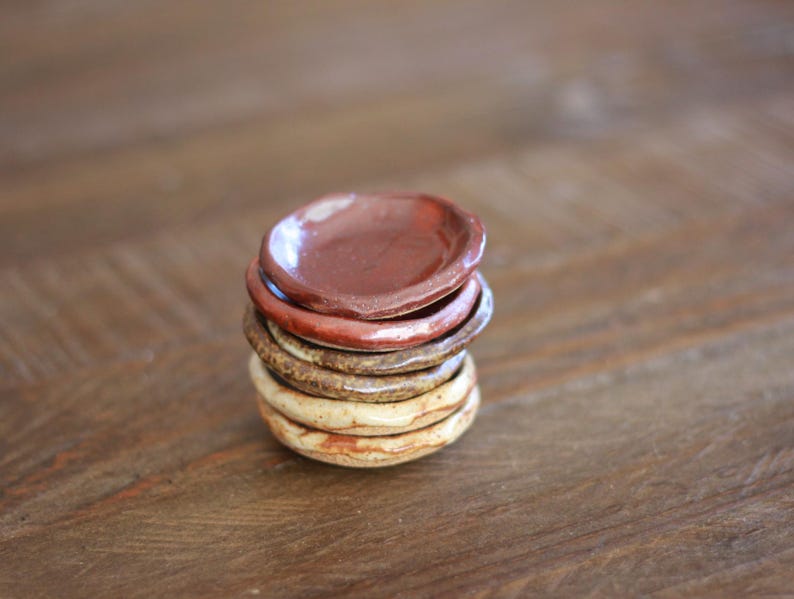 May include: A stack of small, handmade ceramic dishes in varying earth tones. The top dish is a deep reddish-brown, followed by shades of brown and beige. The dishes are stacked on top of each other, creating a rustic and natural aesthetic.
