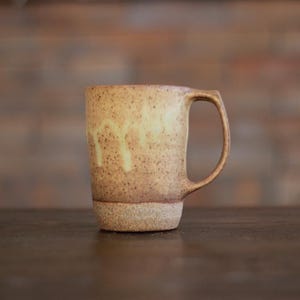 May include: A ceramic mug with a speckled tan and brown finish, featuring a lighter cream-colored glaze dripping down the upper portion. The mug has a cylindrical body, a flat base, and a single curved handle attached to the side.