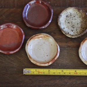 May include: A collection of small, handmade ceramic dishes in various earth-tone colors. The dishes are round with slightly irregular edges. A yellow measuring tape is placed near the dishes, showing inches.
