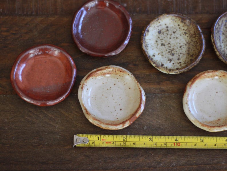 May include: A collection of small, handmade ceramic dishes in various earth-tone colors. The dishes are round with slightly irregular edges. A yellow measuring tape is placed near the dishes, showing inches.
