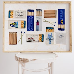 May include: A framed collection of matchboxes with various designs featuring nautical themes and locations on Cape Cod, Massachusetts. The matchboxes are arranged in a grid pattern and include names like "Entering Cape Cod", "Ocean Edge", "Nauset Light", "Coast Guard", "Chatham Pier Fish Market", and "Chatham Bars".