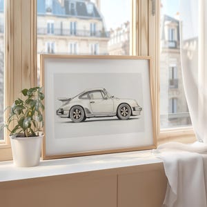 May include: A framed print of a white Porsche 911 car on a light wood windowsill. The car is drawn in a realistic style with a light gray background.