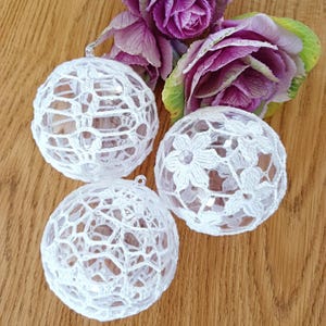 May include: Three white crocheted Christmas ornaments with a delicate floral pattern. The ornaments are made of white crochet thread and are strung on clear glass balls.