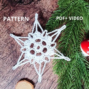 Puede incluir: Adorno de copo de nieve blanco tejido a crochet sobre un fondo de madera con una rama de pino verde. El texto "PATTERN PDF+VIDEO" es visible en la esquina superior derecha de la imagen.