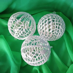 May include: Three white, crocheted sphere ornaments with intricate openwork patterns. Each sphere has a small loop for hanging. The ornaments are displayed against a vibrant green fabric background, creating a visually appealing contrast.