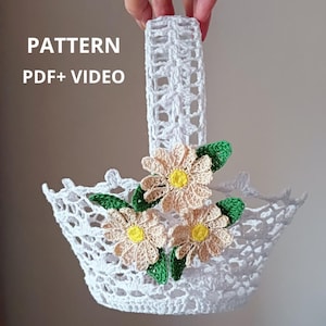 Op de afbeelding: Een witte gehaakte mand met een handvat en drie gehaakte bloemen in crème en gele tinten met groene bladeren. De tekst "PATTERN PDF + VIDEO" is zichtbaar in de linkerbovenhoek van de afbeelding.