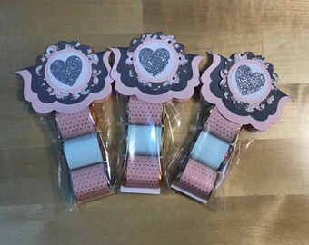 May include: Three small gift bags with pink and gray paper, each containing a small wrapped gift. The bags are decorated with a heart-shaped design with a silver glitter heart in the center.
