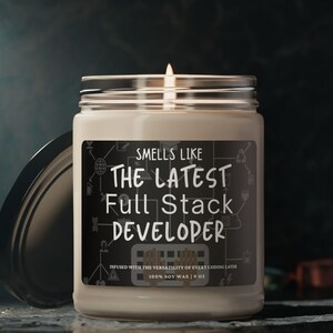 May include: A white candle with a black label that reads "Smells Like The Latest Full Stack Developer". The label is decorated with a circuit board design. The candle is in a glass jar with a black lid.
