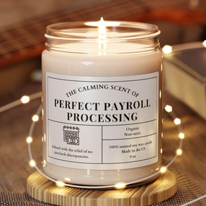 May include: A white candle with a brown label that reads "The Calming Scent of Perfect Payroll Processing". The label also includes the text "Organic, Non-toxic, 100% natural soy wax candle, Made in the US, 9 oz".