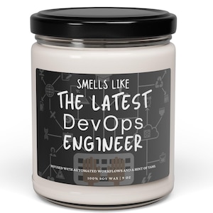 May include: A white candle jar with a black lid and a label that reads "Smells Like The Latest DevOps Engineer" with a graphic of computer icons. The label also says "Infused with automated workflows and a hint of tame. 100% Soy Wax | 9 oz."