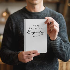 May include: A white notebook with the text "Very Important Engineering stuff" in black script is held in front of a person. The notebook is rectangular and has a clean, minimalist design. The person is wearing a dark gray sweater.