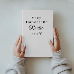May include: A white notebook with the text "Very Important Realtor stuff" in black script and sans-serif fonts. The notebook is held by two hands, with rings on the fingers, against a light background.