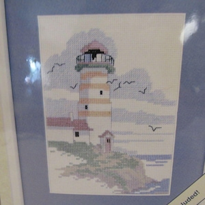 Puede incluir: Una bordado de punto de cruz que representa un faro con una torre de rayas blancas y naranjas, un techo rojo y una pequeña casa a su lado. El faro se encuentra en un acantilado con vistas al mar con agua azul y olas blancas. El cielo es azul claro con nubes blancas y pájaros volando.