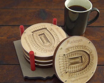 May include: Wooden coasters with a stadium design, stacked on a gray base. The coasters are engraved with the word "Buckeyes" and the name "Tammy Alderman". A mug of coffee sits nearby, with red posts holding the coasters.