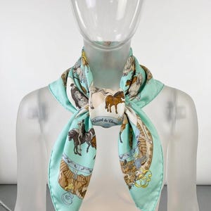 May include: A silk scarf with a light blue border and a central design featuring horses and riders in various poses. The scarf has a cream-colored background with brown and black horses. The text "Avant du Chasse" is visible.