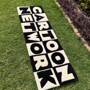 Könnte beinhalten: Ein rechteckiger schwarz-weißer Teppich mit den Worten "CARTOON NETWORK" in großen, fetten Buchstaben. Der Teppich liegt auf einer grünen Grasfläche, wobei der Text in einem Blockbuchstabenstil gehalten ist.