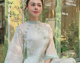 May include: A woman wearing a light green, floral patterned dress with a white headband. The dress has a mandarin collar and a tassel detail. The woman is sitting in front of a green door with a sign that says "indoor coffee" and "come in, it's really happy inside."