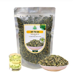 May include: A resealable bag of dried green tea leaves labeled "TRÀ TÂM SEN" with the text "Tinh Hoa Thảo Mộc" and "Cải Thiện Các Ngủ Thanh Lạc Thể" in Vietnamese. The bag is sitting on a white surface with a small bowl of the tea leaves and a glass of tea.