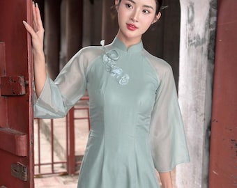 May include: A woman wearing a light green dress with a high collar and sheer sleeves. The dress has a floral embellishment on the chest. She is standing in a doorway with a woven bag in her hand.