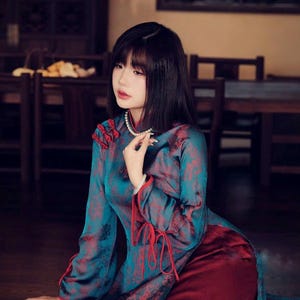 May include: A woman wearing a teal and burgundy traditional dress with a floral pattern. The dress has long sleeves with red ties at the cuffs and a high collar. She is wearing a pearl necklace.