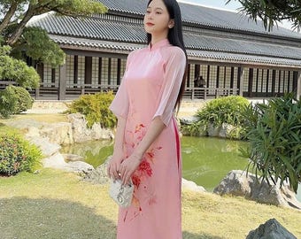 May include: A woman wearing a pink and white long-sleeved dress with floral print and a pink skirt. She is holding a white handbag and standing in front of a pond and a building with a tiled roof.