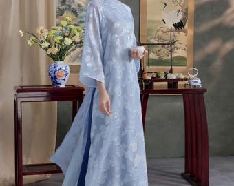 May include: A light blue, long-sleeved dress with floral embroidery. The dress has a high collar and a flowing skirt, paired with dark blue pants. The woman is holding a small white cup. The background includes traditional artwork and dark wood furniture.