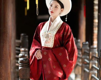May include: A person wearing a traditional red and white Asian-style dress and hat. The dress has floral embroidery and a patterned design. The background includes wooden structures and hanging lanterns.