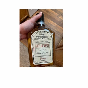 May include: A 750ml glass bottle filled with amber liquid, featuring a custom label that reads: Larry, We would be honored, if you did the honors. Will you marry us? SAT 12/18/21, Requested by Karrie & Dilan, Emory Texas. The bottle has a black cap.