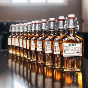 May include: A row of amber glass bottles with personalized labels for a wedding. The labels are white with black text and include the names of the groomsmen, the date of the wedding, and the words "Groom" and "Kat".