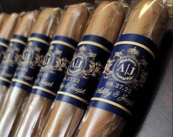 May include: Several cigars are displayed, each wrapped in clear cellophane. The cigars have a brown hue and are adorned with a navy blue band featuring gold lettering and a decorative emblem. The band includes the initials "A/J" and the date "08.27.22".